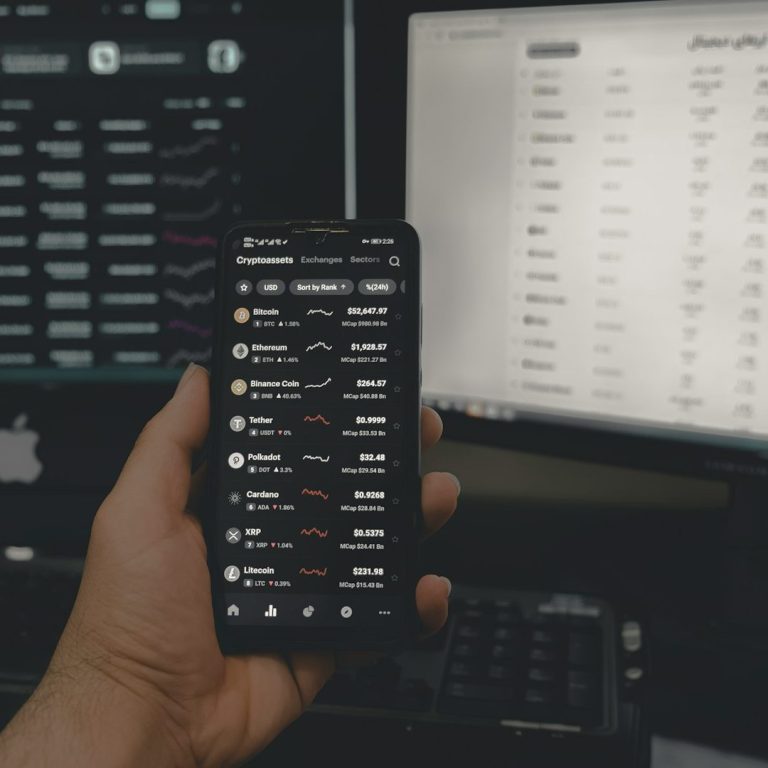 Hand hält ein Smartphone mit Finanzdaten, im Hintergrund mehrere Computerbildschirme.