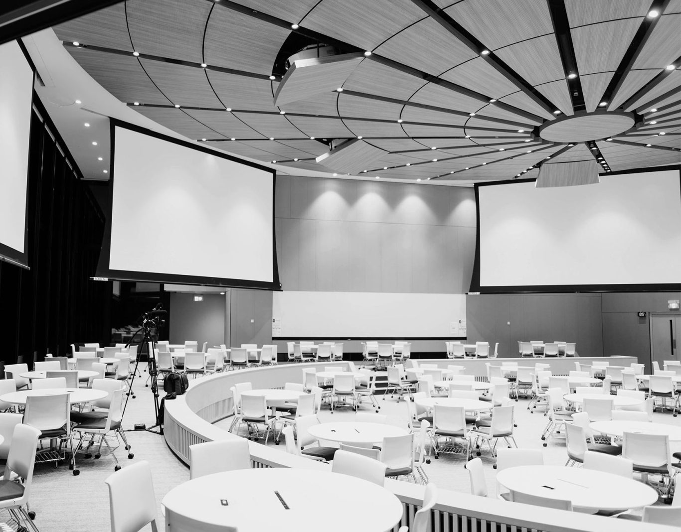 Rundsaal mit Tischen und Stuhlreihen, Leinwände an der Decke montiert.