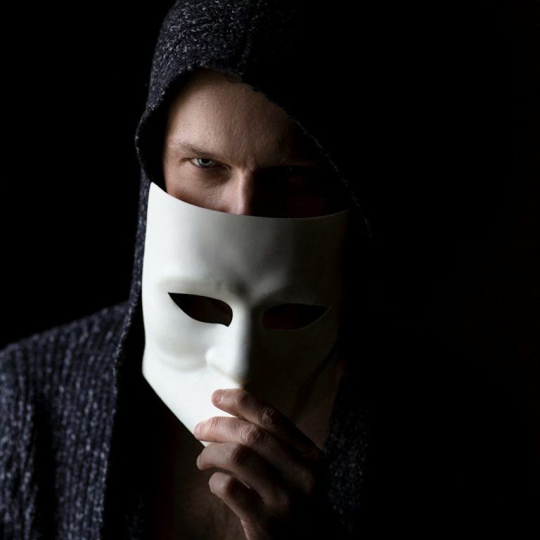 Person mit dunkler Kapuzenjacke hält eine weiße Maske vor dem Gesicht.