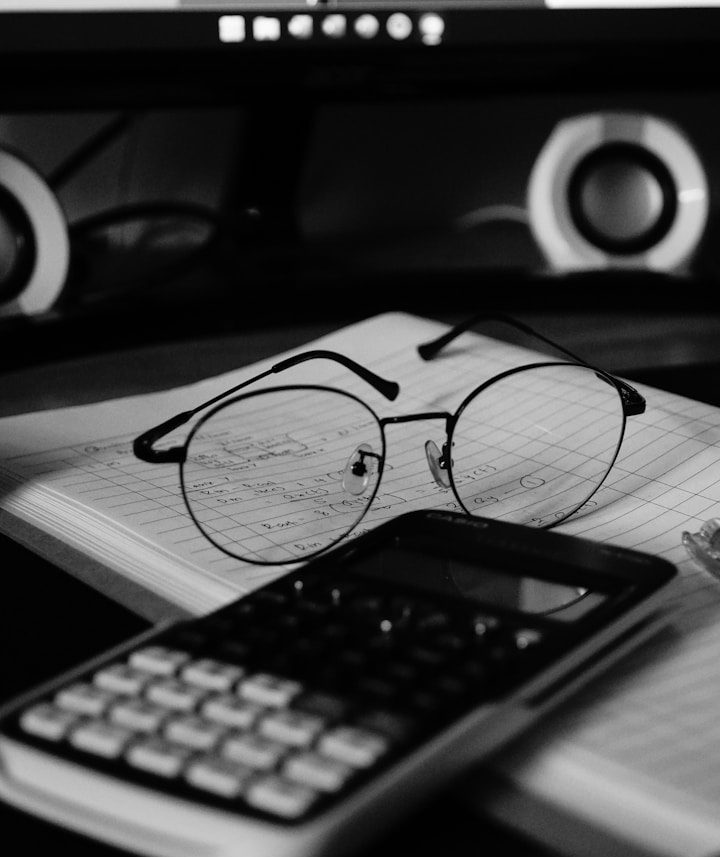 Brille, Taschenrechner und Notizbuch liegen auf einem Tisch vor einem Computer.
